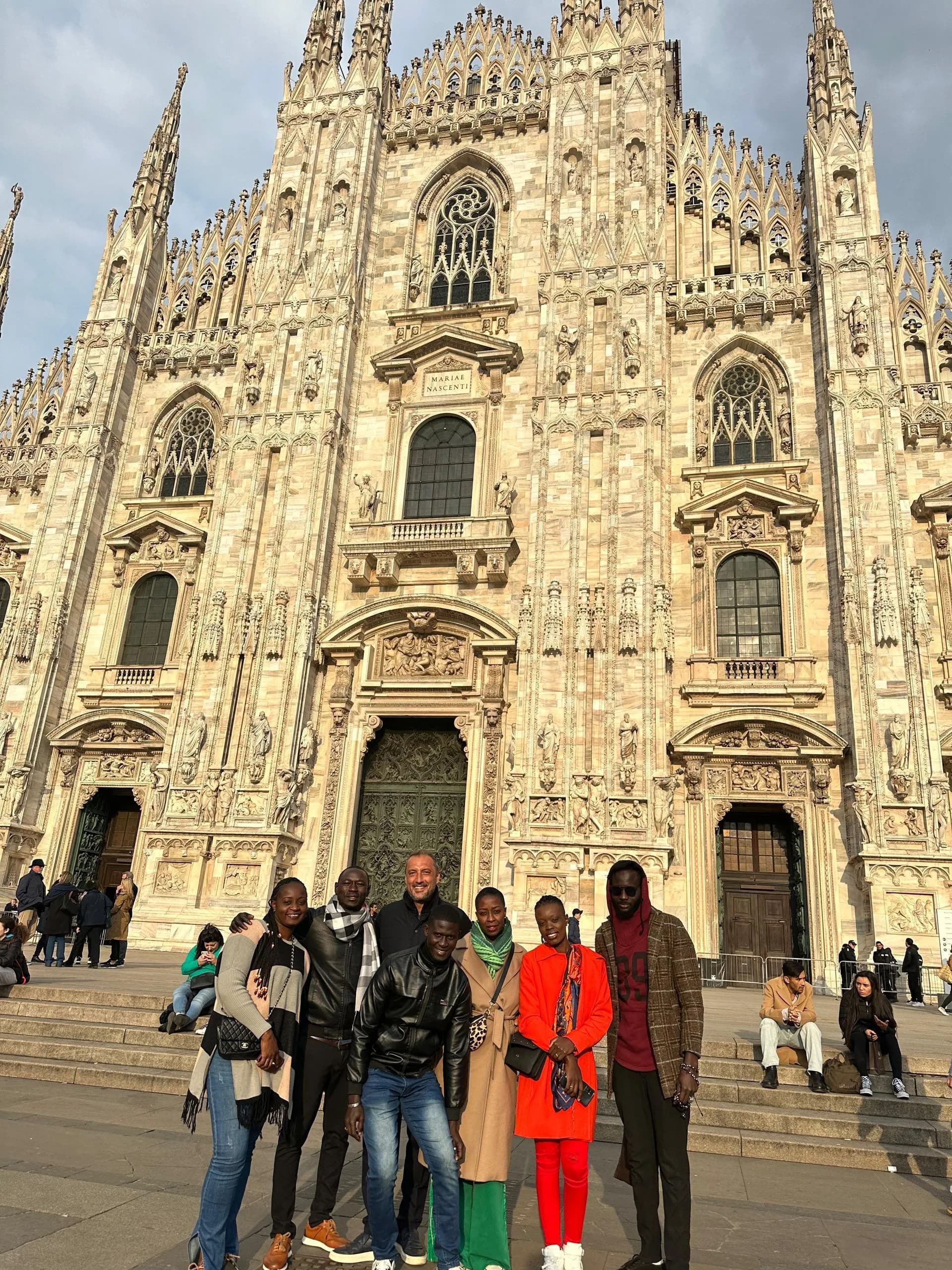 Des photos de groupe sont prises devant la cathédrale de Milan, richement ornée. Des personnes se tiennent debout ou assises.