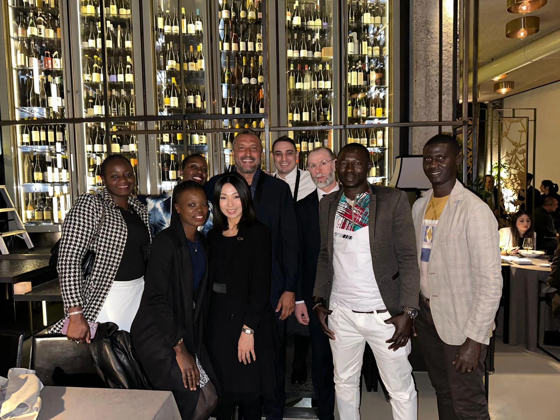 Un groupe de personnes pose pour une photo dans un restaurant ; des bouteilles de vin sont visibles derrière eux.